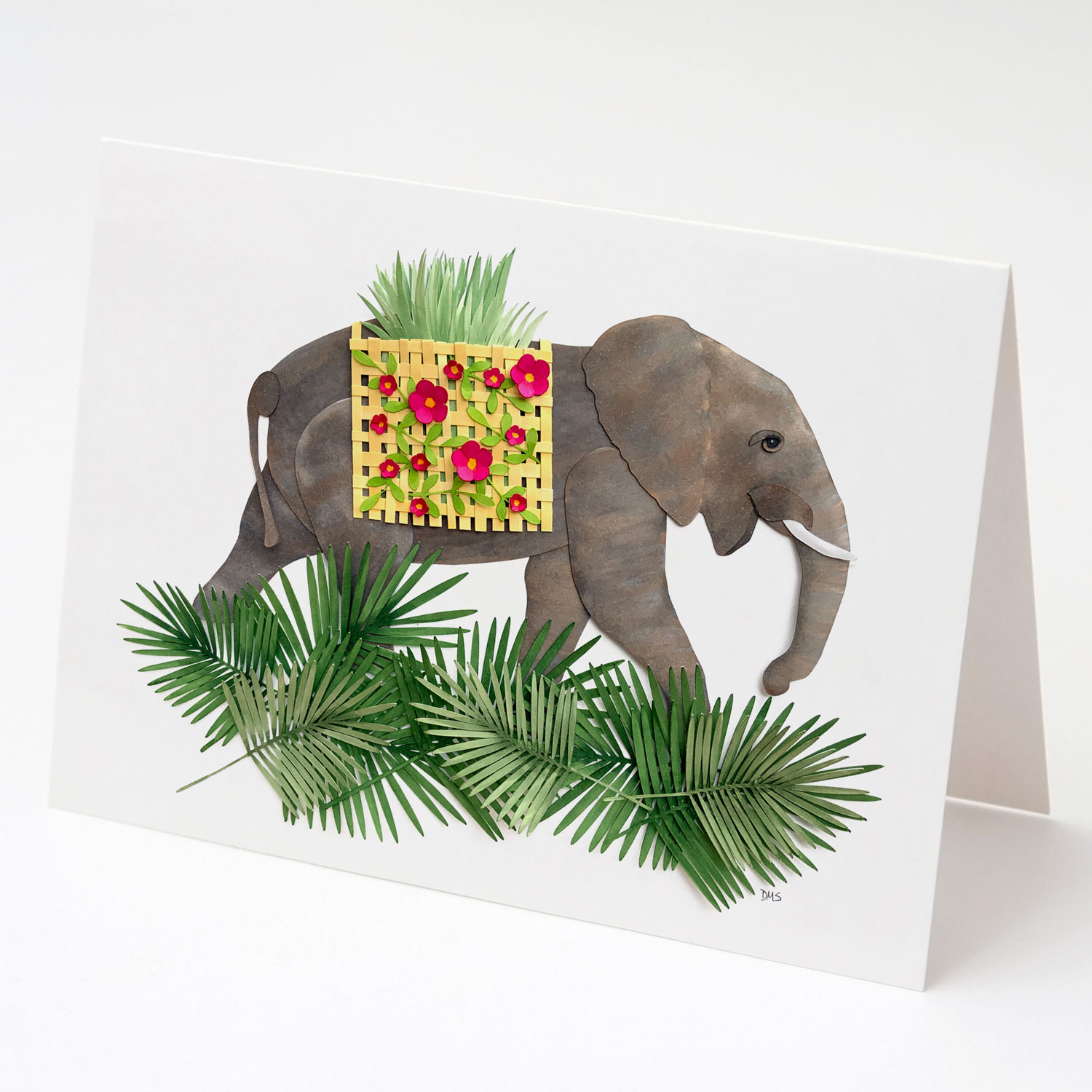 A card depicting a happy, walking paper elephant carrying a woven basket of dried grasses. The basket is adorned with pink flowers and there are palm fronds at the elephant's feet.