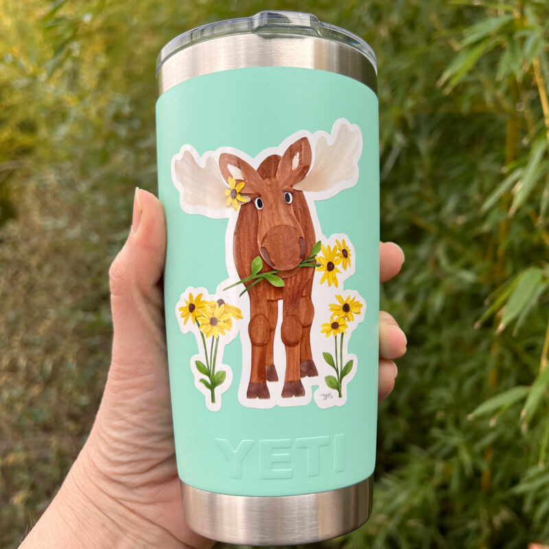 A moose sticker, depicting a cute moose surrounded by black-eyed Susan flowers, on an aqua colored Yeti mug.