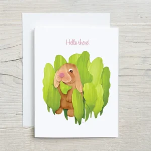 Paper sculpture greeting card showing a lop-eared bunny peeking out from behind a mound of green leaves with Hello There printed at the top